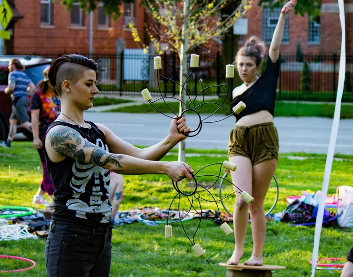 A flow artist practicing with two flower-shaped props while another balances nearby in Palmer Square Park